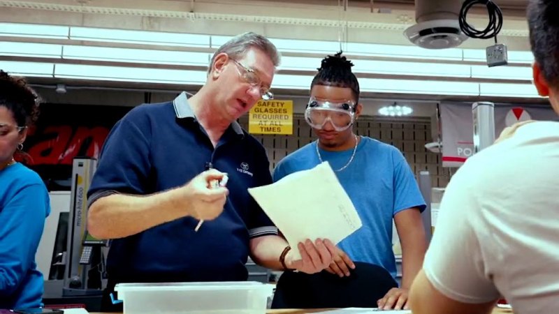 An instructor holding a sheet of paper talks to a student wearing safety glasses.